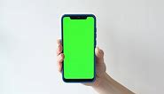 Hand of young woman holding a smartphone with vertical green chroma key screen on gray backdrop. 4k template. Girl hands keeps cell phone with green mockup screen background in front of the camera