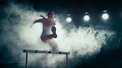 Strong Female Athlete is Running Towards an Obstacle, Jumping Over the Barrier at High Speed while Sprinting. Cinematic Dark Studio Sports Footage with Super Slow Motion Speed Ramp and Smoke Effects