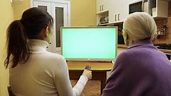 Family watching TV. Green screen. A young woman and a grandmother are sitting at home in the kitchen and watching TV. The woman presses the buttons on the TV remote control.