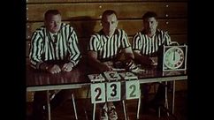 1970s: Referees sitting at table at wrestling match change score card to OT. Ref flips disc like coin. Two wrestlers face off on hands and knees.