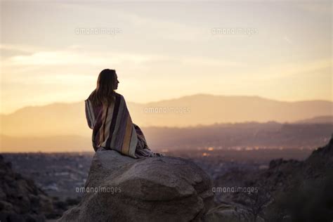 Rear View Of Woman Wrapped In Blanket Sitting On Rock Against Sky