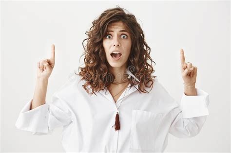 surprised puzzled european female with curly hair pointing up with both
