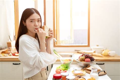 Asian Young Woman Eating Sandwich For Breakfast In The Kitchen Stock