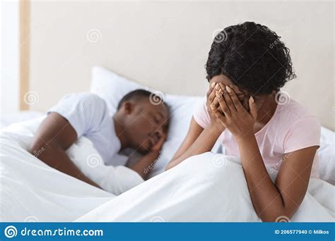 Crying African Woman Sitting In Bed While Her Husband Sleeping Stock