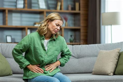 Unhealthy Senior Woman Sitting On Sofa At Home Holding Her Stomach