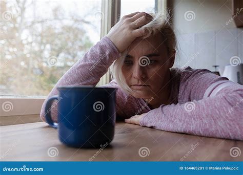 Blonde Woman Sitting At The Kitchen Table Covering Her Face With Her