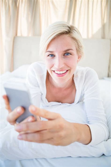 smiling blonde woman lying on the bed and texting with her mobile phone