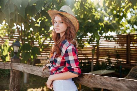 cute smiling redhead cowgirl in hat leaning on ranch fence