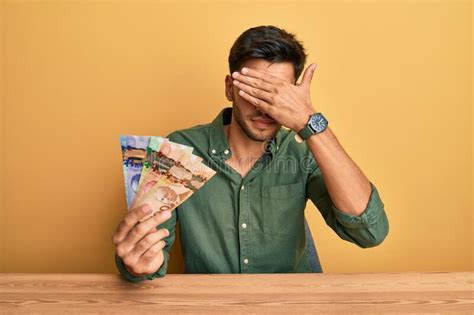 young handsome man holding canadian dollars covering eyes  hand