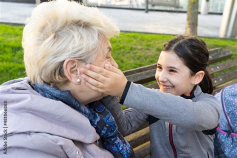 Smiling Granddaughter Caresses Senior Blonde Woman Sitting On Park