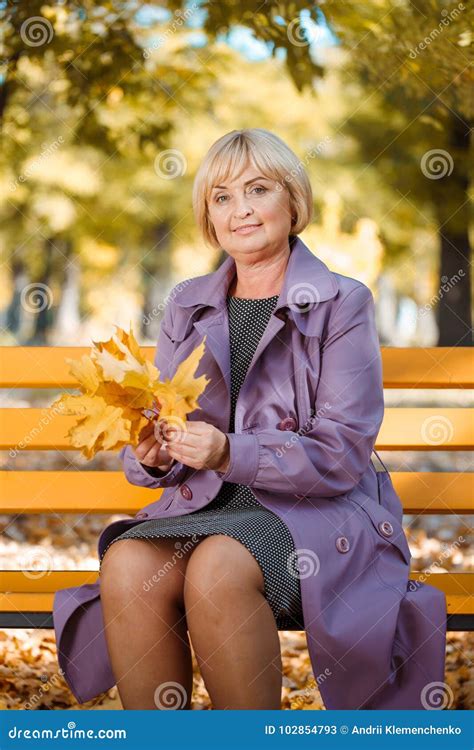 A Mature Woman Sits On A Bench In The Park And Holds Autumn Leaves My