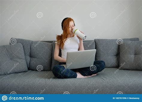 Redhead Girl In Pajamas Sits On A Sofa With A Laptop On Her Folded Legs
