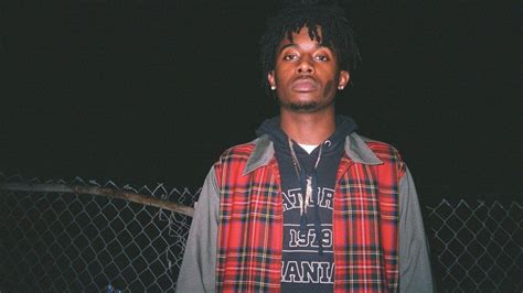 playboi carti  wearing red striped coat standing  fence black background wearing gold chains