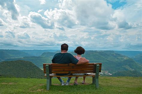 couple sitting  brown wooden bench  mountains covered  grasses  blue cloudy sky