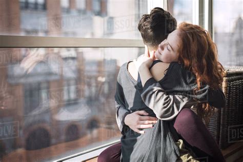 Lesbian Couple Embracing While Sitting On Window Sill At Home Stock