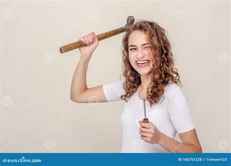 Curly Cheerful Brunette Girl In Denim Overalls Holds A Hammer In Her