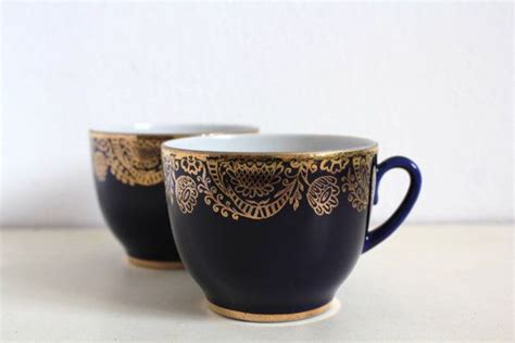 Two Black And Gold Coffee Mugs Sitting Next To Each Other On A White