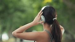 Happy asian woman adjusting wireless headphones before starting jogging and listening to music on running route surrounded by nature. Slow motion.