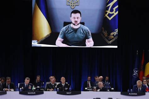 Zelensky wearing a green shirt speaks in a dimly-lit room. An out-of-focus Christmas tree is in the