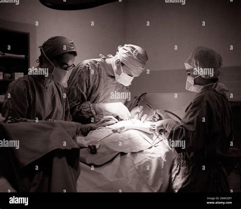 1960s MAN DOCTOR SURGEON AND TWO WOMEN ATTENDANT SURGICAL NURSES ASSISTING PERFORMING A SURGERY ...
