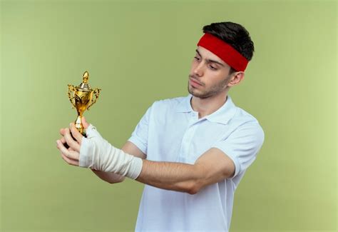 Young sporty man in headband holding his trophy looking at it with ...
