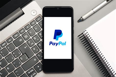 Paypal logo on black screen of smartphone with laptop and notebook ...