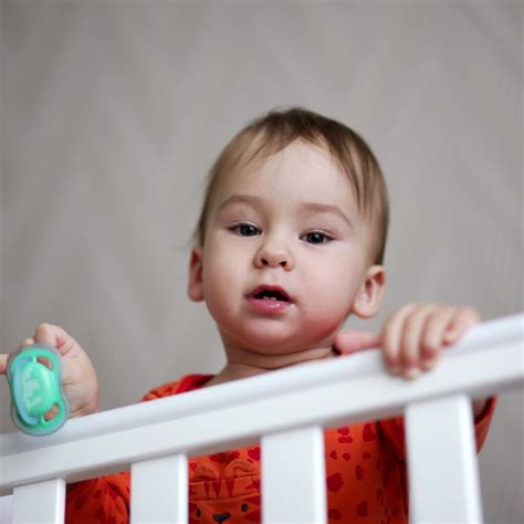 Cute one-year-old child standing in the crib and holding a pacifier in ...