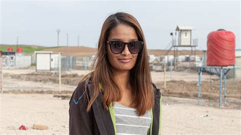 Shamima Begum leaning against a door frame. She has long dyed brown hair and is wearing sunglasses with a light grey top underneath a sweatshirt jacket