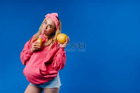 蓝色背景中穿着粉色衣服、一瓶果汁和一个瓜的怀孕女孩高清摄影大图-千库网