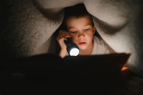 Boy with flashlight reading book under blanket at home | Premium Photo