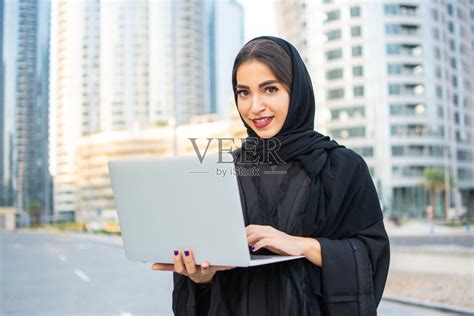 女穆斯林学生在户外穿着阿拉伯长袍拿着笔记本电脑照片摄影图片_ID:353491326-Veer图库