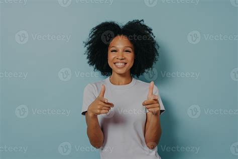 Positive friendly dark skinned lady making finger gun gesture while standing on blue background ...
