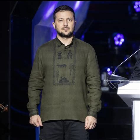 Zelensky wearing a green shirt speaks in a dimly-lit room. An out-of-focus Christmas tree is in the