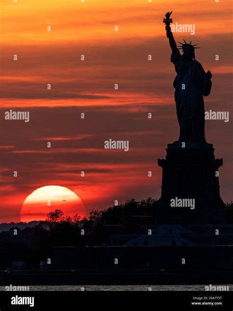 Silhouette of Statue of Liberty with huge setting sun in background and orange red and yellow ...