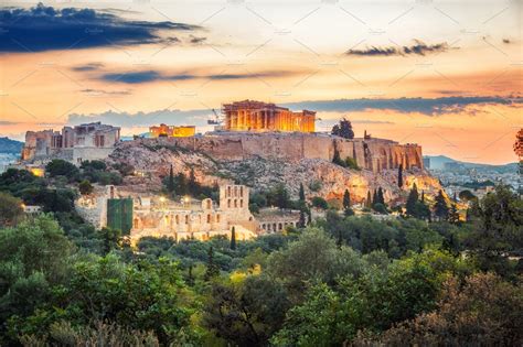 Panoramic sunset view of the Acropolis of Athens, Greece, with the illuminated Parthenon glowing above the ancient hill and lush greenery below.