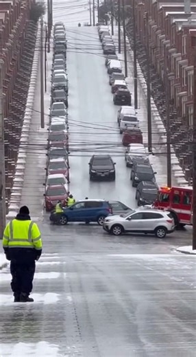 Inattentive Driver Slides Down Icy Hill, Nearly Striking Emergency Crews at Crash Scene Pittsburgh, Pennsylvania. Traffic camera footage shows a steep residential hill coated in ice, with parked cars lining both sides of the street and a collision already blocking the intersection below. Emergency vehicles are positioned across the roadway as responders stand in the street directing traffic and assessing damage. Moments into the clip, a vehicle appears at the top of the hill and begins descendin