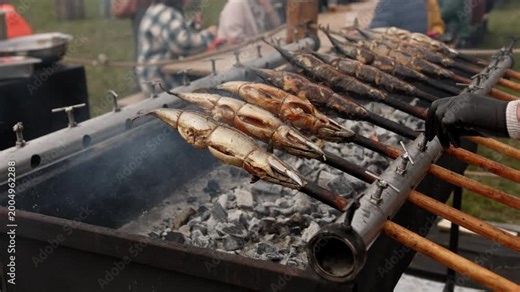 Cook turning fish on a traditional rotisserie grill over charcoal. Street food chef grilling seafood outdoors. Process of preparing roasted fish for meal time.