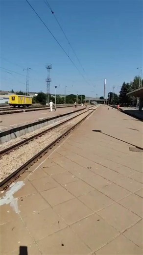 A video shows a train station platform with tracks and a yellow train car in the background. Several people are visible on the platform, walking and standing. The sky is clear and blue, suggesting a sunny day. The platform is paved with square tiles. in Pleven, Bulgaria | Collective Memory