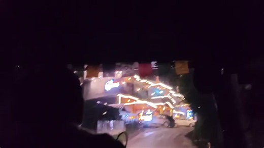 The video shows a point-of-view shot from inside a vehicle at night. The camera is looking forward through the windshield. The silhouette of a person's head and shoulders is visible in the foreground, obscuring the view of the driver. Outside the vehicle, a building is illuminated with decorative lights. Cars are parked in front of the building. The building appears to be a hotel or restaurant with multiple levels and balconies. Prayer flags are hanging from the ceiling of the vehicle. The overa