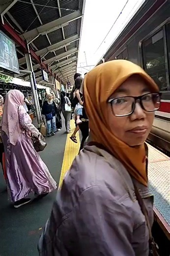The video shows a train platform with several people waiting to board a train. The platform has a yellow safety line along the edge. A train is stopped at the platform, and people are seen boarding it. One woman in a pink dress and headscarf is boarding the train. Another woman in a patterned top and black pants is holding a drink and looking towards the train. A woman in a grey headscarf is standing on the platform. Other people are visible in the background, some appearing to be passengers and