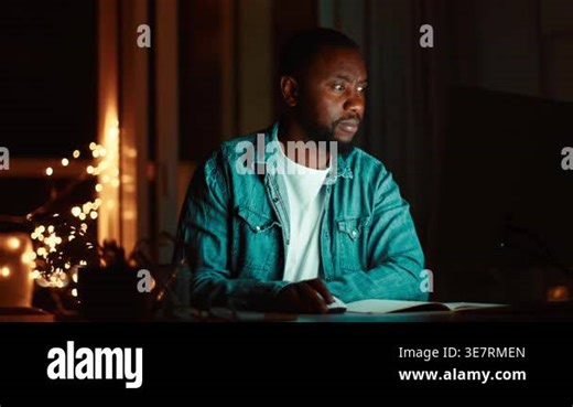 A man with dark skin is focused on his laptop at a desk in his home. Dim lighting and warm fairy lights create a cozy atmosphere. The scene shows concentration and productivity in the evening Stock Video Footage - Alamy