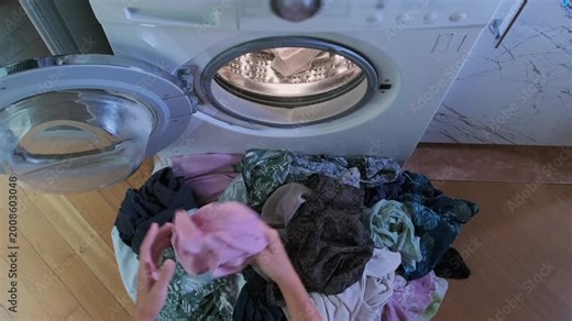 Top view of woman picking up pile of dirty laundry and placing clothes into front loading washing machine, sorting fabrics before wash cycle, everyday household routine indoors.
