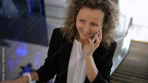 Businesswoman goes up escalator to office, talks on phone and looks at time on watch. Businesswoman is in hurry to get to work. Concept of busy businesswoman in process of active work.