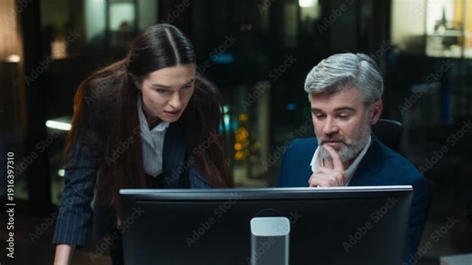 Problem solving moment inside corporate office. Caucasian man sitting at monitor thinking. Female colleague leaning forward discussing data. Team reviewing screen together. Night office.