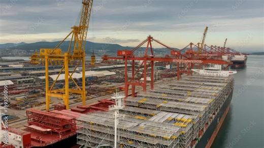 Cranes loading containers on cargo ships at a bustling port with calm waters and distant mountains under a cloudy sky