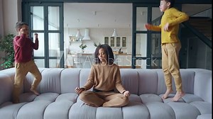 Boys make noise while jumping on the couch in the side of their exhausted mother. Tired African American woman shakes her head, closes her eyes, begins to meditate in lotus position. Mom performs yoga