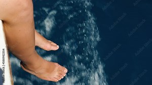 A side view of a woman's bare feet dangling from the side of the ship. A woman's feet against the glistening, frothy white waves of the sea. Cruising. In slow motion