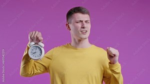 Woken up by alarm clock sleepy young man holding it in hand. Pink background