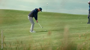 Male golfer goes up and down for par using wedge for chip shot onto green with flag stick blowing in the breeze on Ireland links golf course