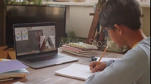 Little Asian boy writing in copybook at table and listening to female school teacher during online lesson via video call on laptop while studying at home during coronavirus quarantine
